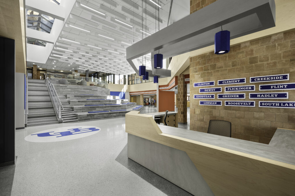 Interior view of The Landing inside UB’s Ellicott Complex on the North Campus, showing a bright student commons with terrazzo flooring, a large UB logo, tiered seating steps, skylights, and a modern reception desk. Blue pendant lights, residence hall signage, and open gathering spaces highlight the contemporary student housing and campus design.