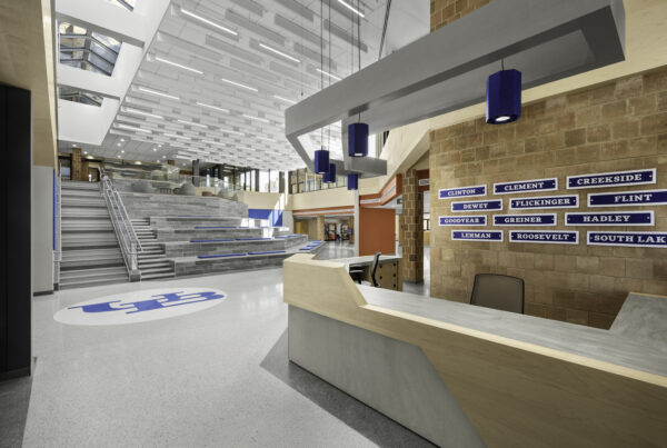 Interior view of The Landing inside UB’s Ellicott Complex on the North Campus, showing a bright student commons with terrazzo flooring, a large UB logo, tiered seating steps, skylights, and a modern reception desk. Blue pendant lights, residence hall signage, and open gathering spaces highlight the contemporary student housing and campus design.